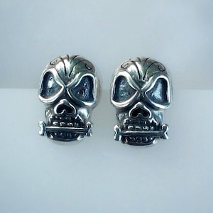 Puede incluir: Un par de pendientes de calavera de plata con un detalle de hueso. Las calaveras tienen detalles precisos, incluidos los ojos, la nariz y la boca.