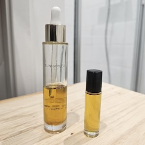 Puede incluir: Dos botellas de aceite esencial de color ámbar. La botella más grande es de vidrio transparente con una tapa dorada y un gotero. La botella más pequeña es una botella de vidrio ámbar oscuro con tapa de rodillo negra. Ambas botellas tienen la etiqueta "SunnaMusk London" y "Fine Oil" impresa.