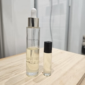 May include: Two glass bottles of essential oil. The larger bottle is clear with a silver cap and a dropper. It says "Sunnamusk London Fine Oil" on the label. The smaller bottle is clear with a black cap and a roller ball. Both bottles are on a light wood surface.