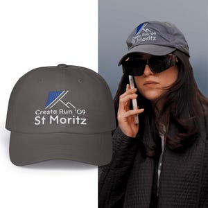 Puede incluir: Una gorra de béisbol gris oscuro con bordado blanco que dice "Cresta Run '09 St. Moritz".