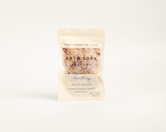 May include: A small, white pouch with a resealable zipper contains a pink and orange bath soak. The pouch has the text "BATH SOAK RESTORE" and "Aromatherapy BATH SALTS" printed on it. The text "LAVENDER, ROSEMARY & CARDAMOM ESSENTIAL OILS" is also printed on the pouch. The pouch is labeled "ARTHUR BETSY".