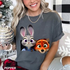 Pu&ograve; includere: T-shirt grigio scuro con una grafica di Judy Hopps e Nick Wilde di Zootropolis. I personaggi dei cartoni animati sono in un design a forma di tasca. La parola "PEPPER" &egrave; stampata in basso sulla maglietta.