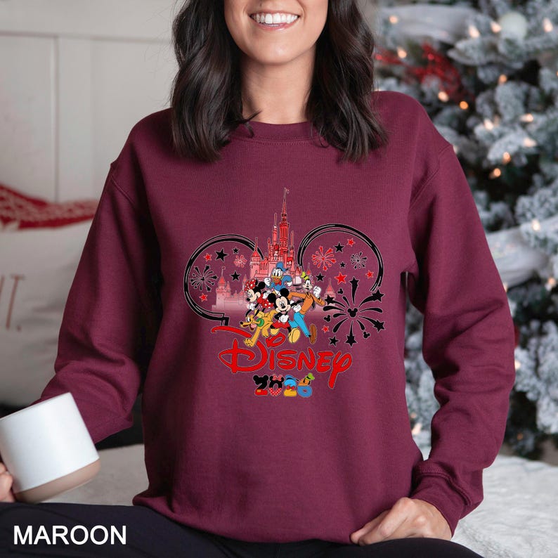 May include: Maroon sweatshirt featuring a Disney design. The graphic includes Disney characters, a castle, fireworks, and the word "Disney" in red. The design is set within Mickey Mouse ear shapes, with the year "2020" below.