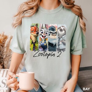 Puede incluir: Camiseta verde claro con un gr&aacute;fico colorido de personajes de la pel&iacute;cula Zootopia. El dise&ntilde;o incluye a Nick Wilde, Judy Hopps, el Jefe Bogo y Flash, con el texto "Zootopia 2" debajo.