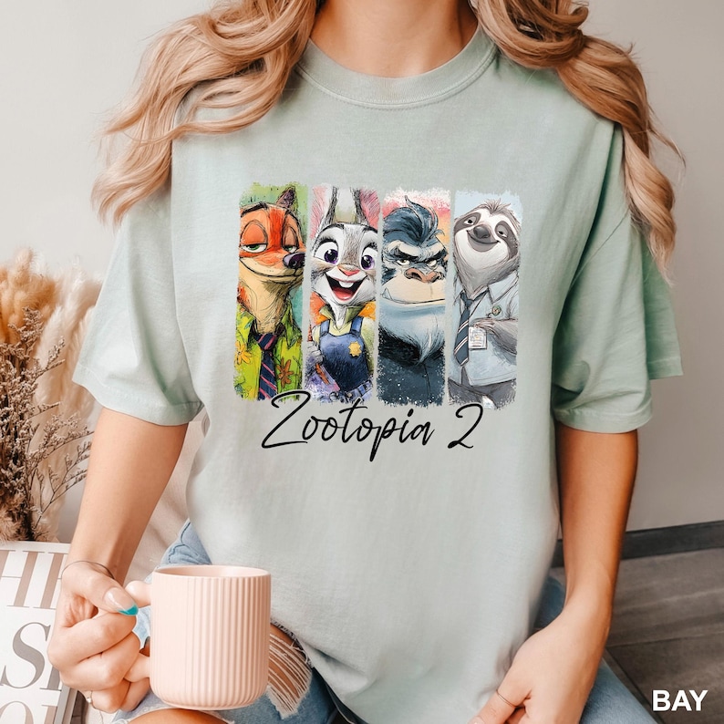 Puede incluir: Camiseta verde claro con un gr&aacute;fico colorido de personajes de la pel&iacute;cula Zootopia. El dise&ntilde;o incluye a Nick Wilde, Judy Hopps, el Jefe Bogo y Flash, con el texto "Zootopia 2" debajo.