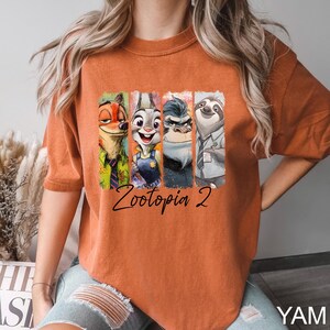 Puede incluir: Camiseta de color naranja quemado con un gr&aacute;fico colorido de personajes de la pel&iacute;cula "Zootopia 2". El dise&ntilde;o incluye a Nick Wilde, Judy Hopps, el Jefe Bogo y Flash. Las palabras "Zootopia 2" est&aacute;n escritas en cursiva debajo de los personajes.