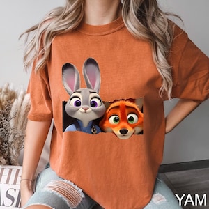 Pu&ograve; includere: T-shirt arancione bruciato con una grafica di Judy Hopps e Nick Wilde di Zootopia. I personaggi dei cartoni animati sono in un ritaglio rettangolare. La maglietta ha maniche corte e una vestibilit&agrave; ampia. Il testo "YAM" &egrave; in basso a destra.