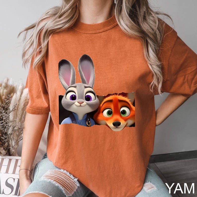Pu&ograve; includere: T-shirt arancione bruciato con una grafica di Judy Hopps e Nick Wilde di Zootopia. I personaggi dei cartoni animati sono in un ritaglio rettangolare. La maglietta ha maniche corte e una vestibilit&agrave; ampia. Il testo "YAM" &egrave; in basso a destra.