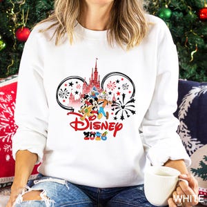 May include: White sweatshirt featuring a Disney design with Mickey Mouse ears, a castle, and various Disney characters. The word "Disney" is in red script, with "2020" below. Black and red fireworks and stars complete the design.