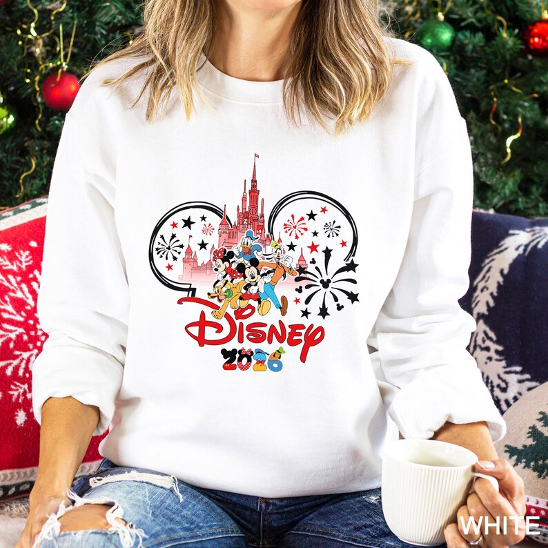 May include: White sweatshirt featuring a Disney design with Mickey Mouse ears, a castle, and various Disney characters. The word "Disney" is in red script, with "2020" below. Black and red fireworks and stars complete the design.