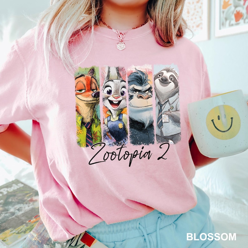 Puede incluir: Sudadera rosa claro con un estampado colorido de personajes de la pel&iacute;cula "Zootopia 2". El dise&ntilde;o incluye a Nick Wilde, Judy Hopps, el jefe Bogo y Flash. Una taza blanca con una carita sonriente amarilla se sostiene en la mano.