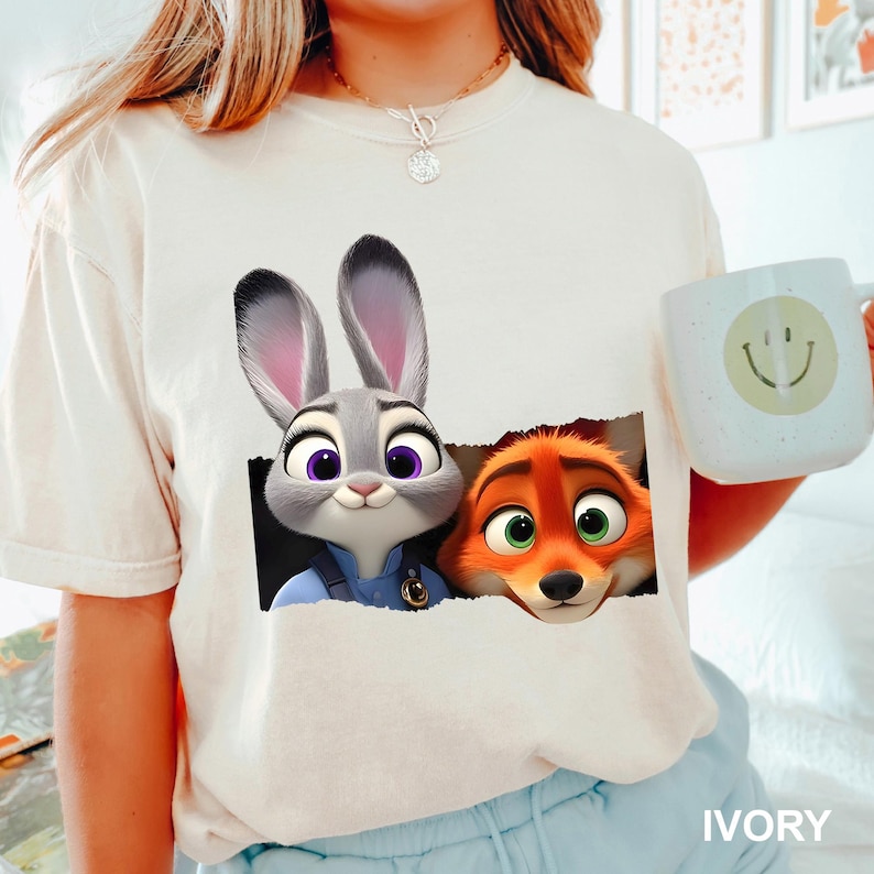 Pu&ograve; includere: T-shirt color avorio con una grafica di Judy Hopps e Nick Wilde di Zootopia. Judy, un coniglio grigio, e Nick, una volpe arancione, sono raffigurati mentre sbucano da un rettangolo nero. Nella foto &egrave; presente una tazza azzurra con una faccina sorridente.