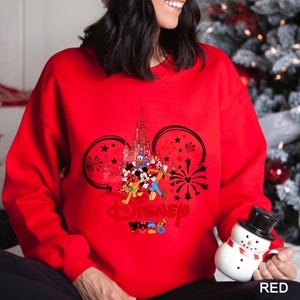 May include: A red sweatshirt featuring a Disney design with Mickey Mouse, Donald Duck, and other characters. The design includes a castle, fireworks, and the word "Disney" in red. The year "2023" is also included in the design.