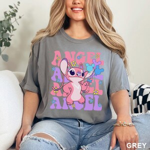 May include: Grey t-shirt featuring the word "ANGEL" in pink and purple retro font, with a pink cartoon character holding blue Mickey Mouse balloons. The shirt is adorned with small flower graphics.
