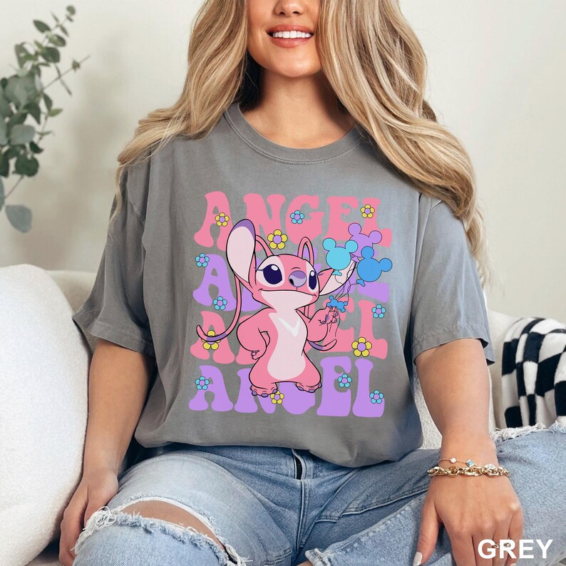May include: Grey t-shirt featuring the word "ANGEL" in pink and purple retro font, with a pink cartoon character holding blue Mickey Mouse balloons. The shirt is adorned with small flower graphics.