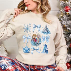 May include: Sand-colored sweatshirt featuring a design with blue bows, snowflakes, a castle, and images of Anna, Elsa, and Olaf from the movie Frozen. The word "SAND" is printed at the bottom.