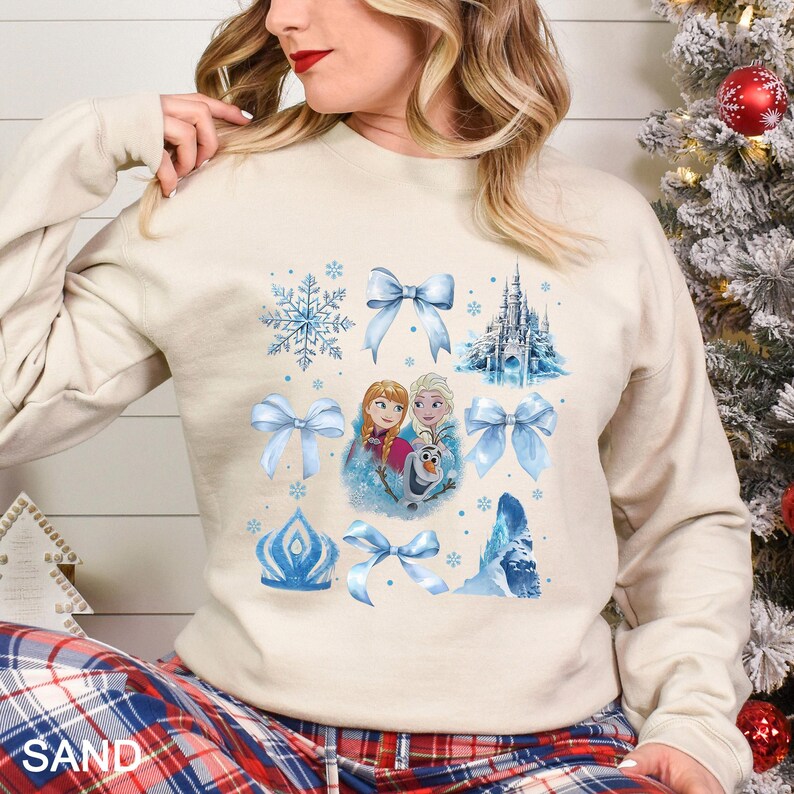 May include: Sand-colored sweatshirt featuring a design with blue bows, snowflakes, a castle, and images of Anna, Elsa, and Olaf from the movie Frozen. The word "SAND" is printed at the bottom.