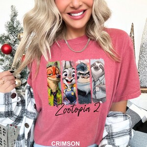 Puede incluir: Camiseta de color carmes&iacute; con los personajes de Zootopia. El dise&ntilde;o incluye a Nick Wilde, Judy Hopps, el jefe Bogo y Flash, con el texto "Zootopia 2" en letra cursiva. La palabra "CRIMSON" est&aacute; impresa en la parte inferior.
