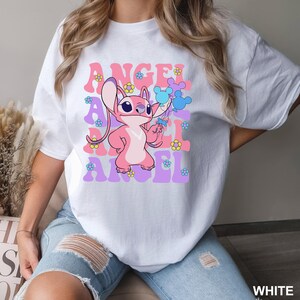 May include: White t-shirt featuring a pink cartoon character holding blue balloons and flowers. The word "ANGEL" is printed in pink and purple, with flower accents. The shirt is a casual style.