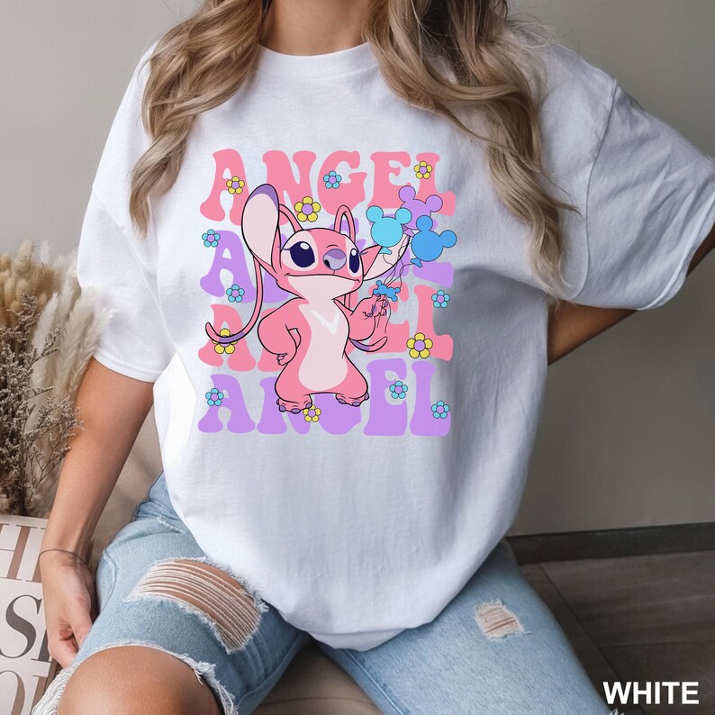 May include: White t-shirt featuring a pink cartoon character holding blue balloons and flowers. The word "ANGEL" is printed in pink and purple, with flower accents. The shirt is a casual style.