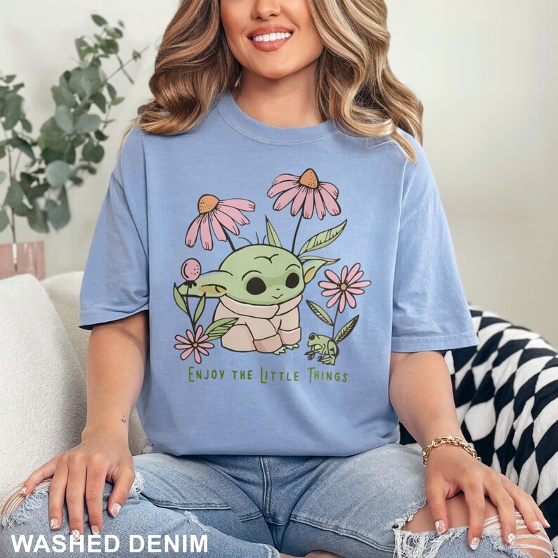 Puede incluir: Camiseta azul claro con un gr&aacute;fico de un personaje verde parecido a un extraterrestre rodeado de flores rosas y el texto "Enjoy the Little Things". La camiseta es de un material suave.