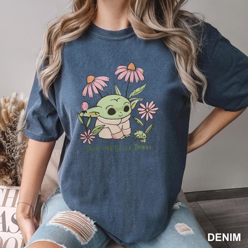 Puede incluir: Camiseta azul denim con un gr&aacute;fico de un personaje verde parecido a un extraterrestre rodeado de flores rosas y el texto "Enjoy the Little Things". La camiseta es de corte holgado.