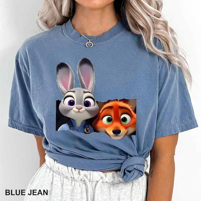 Pu&ograve; includere: T-shirt blu jeans con una grafica di Judy Hopps e Nick Wilde di Zootopia. I personaggi sono in un design rettangolare. Il testo "BLUE JEAN" &egrave; nella parte inferiore della maglietta.