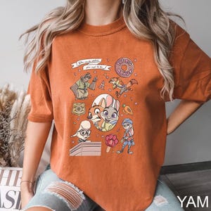 May include: Burnt orange t-shirt featuring a colorful graphic of characters from the animated movie Zootopia. The design includes the text "The Big Donut" and other phrases. The shirt is a comfortable, casual style.