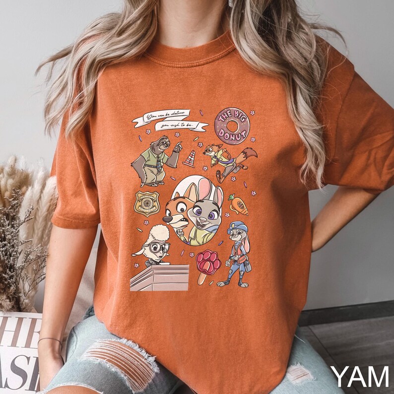 May include: Burnt orange t-shirt featuring a colorful graphic of characters from the animated movie Zootopia. The design includes the text "The Big Donut" and other phrases. The shirt is a comfortable, casual style.