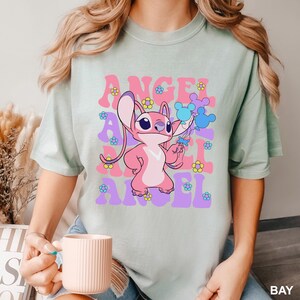 May include: A light green t-shirt featuring a pink cartoon character holding blue balloons. The shirt has the word "ANGEL" in pink and purple, with flower graphics. The shirt is a light green color.