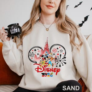 May include: Sand-colored sweatshirt featuring a Disney design with Mickey Mouse, Minnie Mouse, Donald Duck, and Goofy. The design includes a castle, fireworks, and the word "Disney" in red, with "2023" below. The sweatshirt is a comfortable, casual style.