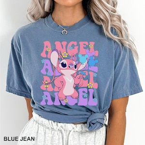 May include: Blue jean colored t-shirt featuring a pink cartoon character holding balloons. The word "ANGEL" is printed in pink and purple retro font with flower accents. The text "BLUE JEAN" is printed at the bottom.