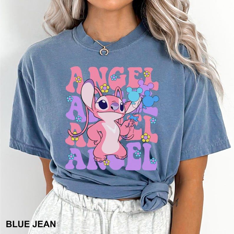 May include: Blue jean colored t-shirt featuring a pink cartoon character holding balloons. The word "ANGEL" is printed in pink and purple retro font with flower accents. The text "BLUE JEAN" is printed at the bottom.