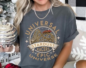 May include: Dark gray t-shirt with a circular graphic featuring a roller coaster and the words "UNIVERSAL", "STARDUST RACERS", and "EPIC UNIVERSE" in gold lettering. The shirt has short sleeves and a crew neck.
