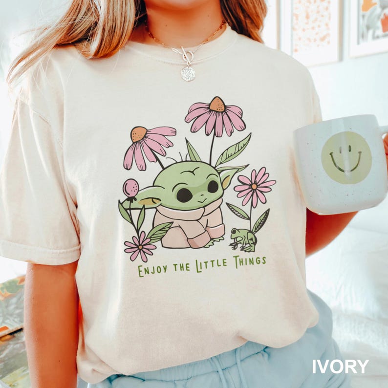 Puede incluir: Camiseta de color marfil con un personaje de dibujos animados verde rodeado de flores rosas y una peque&ntilde;a rana verde. El texto "ENJOY THE LITTLE THINGS" est&aacute; impreso debajo del gr&aacute;fico. Una taza blanca con una carita sonriente est&aacute; en la imagen.