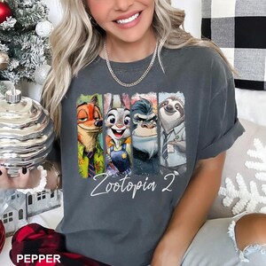 Puede incluir: Camiseta gris oscuro con un estampado de los personajes de Zootopia: Nick Wilde, Judy Hopps, un gorila y Flash el perezoso. El texto "Zootopia 2" est&aacute; escrito en blanco debajo de los personajes.