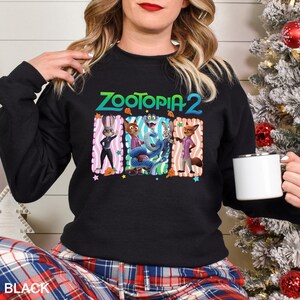 Puede incluir: Sudadera negra de cuello redondo con el texto "Zootopia 2" en verde y azul. El gr&aacute;fico incluye personajes de dibujos animados en marcos coloridos. La palabra "BLACK" est&aacute; impresa en la parte inferior.