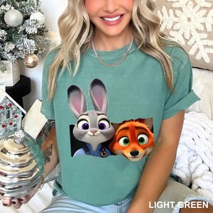 Pu&ograve; includere: T-shirt verde chiaro con Judy Hopps e Nick Wilde di Zootopia. La grafica mostra i personaggi che sbucano da un rettangolo strappato. Il testo "LIGHT GREEN" &egrave; stampato in basso a destra.