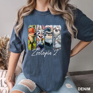 Puede incluir: Una camiseta azul denim con un gr&aacute;fico colorido de personajes de la pel&iacute;cula "Zootopia 2". El gr&aacute;fico incluye a Nick Wilde, Judy Hopps, el Jefe Bogo y Flash. Las palabras "Zootopia 2" est&aacute;n escritas en cursiva.