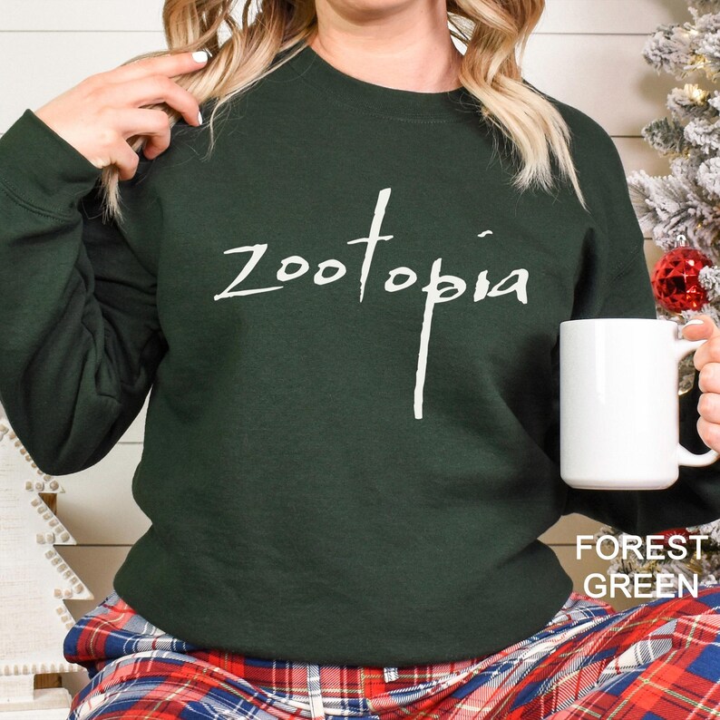 Puede incluir: Sudadera verde bosque con la palabra "zootopia" en escritura blanca. Una taza blanca se sostiene en la mano derecha. Las palabras "FOREST GREEN" est&aacute;n en la esquina inferior derecha de la imagen.