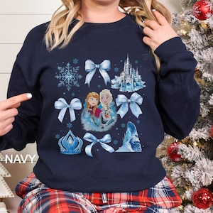 May include: Navy blue sweatshirt featuring a collage of light blue bows, snowflakes, a castle, and images of Anna, Elsa, and Olaf. The word "NAVY" is printed on the left side of the sweatshirt.