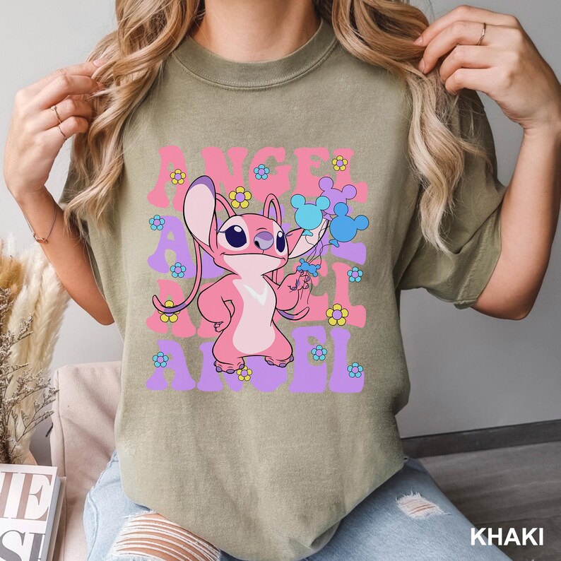May include: Khaki-colored t-shirt featuring a pink cartoon character holding blue and purple shapes. The shirt has the words "Angel" in pink and purple, with flower accents. The bottom right corner of the shirt reads "Khaki".