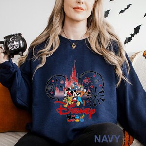 May include: Navy blue sweatshirt featuring a Disney design. The graphic includes a castle, Mickey Mouse, Donald Duck, and other characters. The word "Disney" is in red, and the year "2023" is below it. The sweatshirt is a crew neck style.