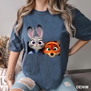 Pu&ograve; includere: T-shirt blu denim con una grafica di Judy Hopps e Nick Wilde di Zootopia. I personaggi dei cartoni animati sono in un design rettangolare. La parola "DENIM" &egrave; stampata in basso a destra.