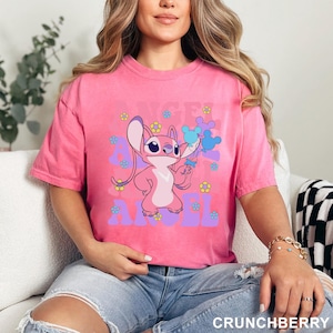 May include: A pink t-shirt featuring a cartoon character holding blue balloons and surrounded by flowers. The text "ANGEL" is printed in purple. The shirt is a solid pink color and has short sleeves.