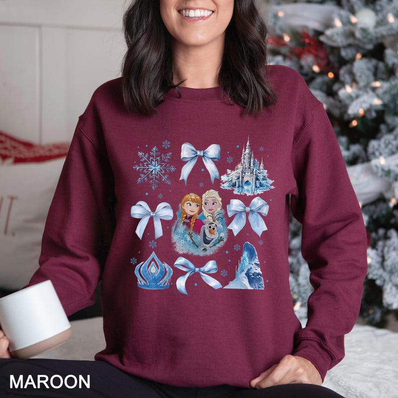 May include: Maroon sweatshirt featuring a design with blue bows, snowflakes, a castle, and images of two girls and a snowman. The word "MAROON" is printed at the bottom of the image.