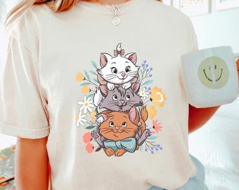 May include: A white t-shirt with a graphic of three cartoon cats stacked on top of each other. The cats are surrounded by flowers and the bottom cat is wearing a blue bow tie. The text "IVORY" is in the bottom right corner of the image.