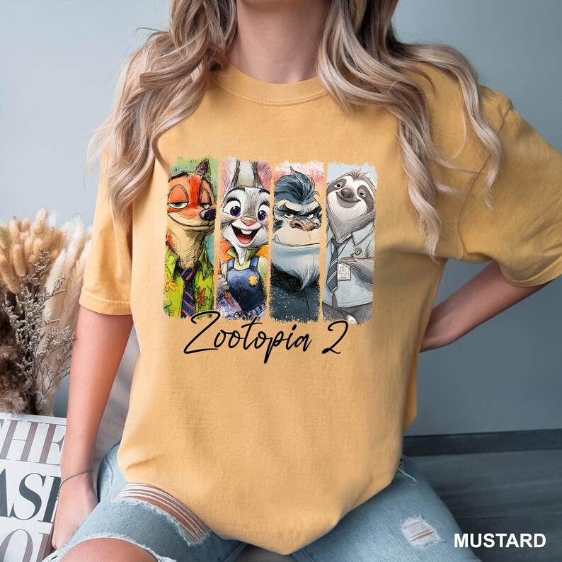 Puede incluir: Camiseta de color mostaza con un gr&aacute;fico colorido de personajes de Zootopia 2. El dise&ntilde;o incluye im&aacute;genes de un zorro, un conejo, un gorila y un perezoso. Las palabras "Zootopia 2" est&aacute;n escritas en cursiva debajo de los personajes.
