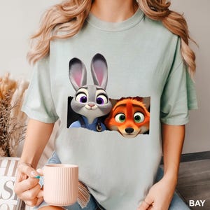 Pu&ograve; includere: T-shirt verde chiaro con una grafica di Judy Hopps e Nick Wilde di Zootropolis. Judy, un coniglio grigio con occhi viola, e Nick, una volpe arancione, sono raffigurati mentre sbucano da un rettangolo strappato. La parola "BAY" &egrave; stampata in bianco in basso a destra.