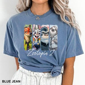 Puede incluir: Camiseta azul vaquero con un gr&aacute;fico colorido de personajes de Zootopia 2. El gr&aacute;fico incluye a Nick Wilde, Judy Hopps, un rinoceronte y Flash el perezoso. El texto "Zootopia 2" est&aacute; escrito en cursiva.